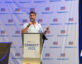A man with short brown hair and a beard is speaking into a microphone while standing behind a podium. The podium has a sign that reads "ClearanceJobs CONNECT 2025" with a small squirrel logo. The background features a large banner with repeated "ClearanceJobs" logos and red, white, and blue flag-like symbols. To the right, there is a display board with various logos and text, partially visible. A dark brown chair is positioned to the left of the podium.