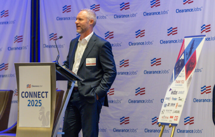 A man in a dark suit and white shirt is standing at a podium with a microphone, speaking at an event. The podium has a sign that reads "ClearanceJobs CONNECT 2025" with a logo of a squirrel wearing a hat. Behind him is a backdrop with the ClearanceJobs logo repeated multiple times. To the right, there is a stand with a large sign displaying various sponsor logos and the text "CONNECT 2025 THANK YOU TO OUR SPONSORS.