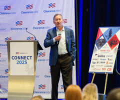 A man in a blue blazer and light blue shirt is speaking into a microphone on stage at a ClearanceJobs event called "CONNECT 2025." He stands next to a podium with the event logo and a signboard displaying sponsor logos, including Leidos, Carahsoft, Peraton, Think, KBR, McDean, Nooks, Anduril, TruVetting, and CGI. The backdrop features the ClearanceJobs logo repeatedly. Audience members are partially visible in the foreground.