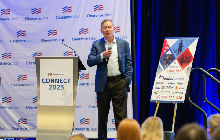 A man in a blue blazer and light blue shirt is speaking into a microphone on stage at a ClearanceJobs event called "CONNECT 2025." He stands next to a podium with the event logo and a signboard displaying sponsor logos, including Leidos, Carahsoft, Peraton, Think, KBR, McDean, Nooks, Anduril, TruVetting, and CGI. The backdrop features the ClearanceJobs logo repeatedly. Audience members are partially visible in the foreground.