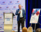 A man in a blue blazer and light blue shirt is speaking into a microphone on stage at a ClearanceJobs event called "CONNECT 2025." He stands next to a podium with the event logo and a signboard displaying sponsor logos, including Leidos, Carahsoft, Peraton, Think, KBR, McDean, Nooks, Anduril, TruVetting, and CGI. The backdrop features the ClearanceJobs logo repeatedly. Audience members are partially visible in the foreground.