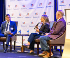 Three people are seated on stage in a panel discussion setting. The person on the left is a man in a blue suit holding a microphone and some papers. The woman in the center is speaking into a microphone, wearing glasses, a dark blazer, and dark pants. The man on the right is wearing a maroon blazer, light blue pants, and brown shoes, holding a microphone in his lap. There is a small round table with two water bottles and some items between the first two people. Behind them is a backdrop with the "ClearanceJobs" logo repeated. A podium on the right side of the image displays a sign that reads "CONNECT 2025.