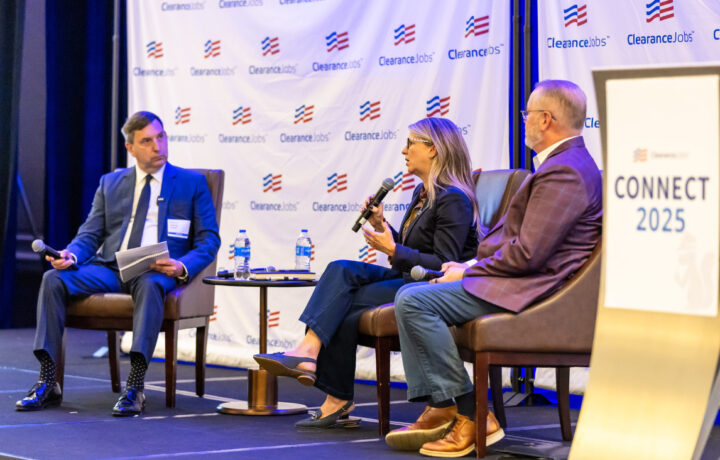Three people are seated on stage in a panel discussion setting. The person on the left is a man in a blue suit holding a microphone and some papers. The woman in the center is speaking into a microphone, wearing glasses, a dark blazer, and dark pants. The man on the right is wearing a maroon blazer, light blue pants, and brown shoes, holding a microphone in his lap. There is a small round table with two water bottles and some items between the first two people. Behind them is a backdrop with the "ClearanceJobs" logo repeated. A podium on the right side of the image displays a sign that reads "CONNECT 2025.