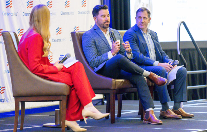 Three people are seated on stage in a panel discussion setting. The person on the left is a woman wearing a bright red suit and beige heels, holding a microphone and some papers. The man in the middle is dressed in a blue checkered blazer, white shirt, and dark pants, speaking into a microphone. The man on the right wears a blue suit with a light blue shirt and brown shoes, holding a microphone and papers. The backdrop features a repeated "ClearanceJobs" logo.