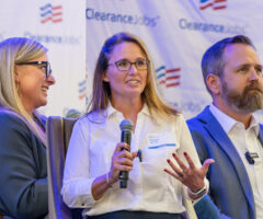 Three people are seated on a panel in front of a backdrop with the "ClearanceJobs" logo. The woman in the center, wearing glasses and a white shirt, is speaking into a microphone and gesturing with her hand. The woman on the left, also wearing glasses and a dark blazer, is smiling and looking toward the center speaker. The man on the right, with a beard and wearing a blue suit jacket, is looking forward attentively. All three have name tags and microphones clipped to their clothing.