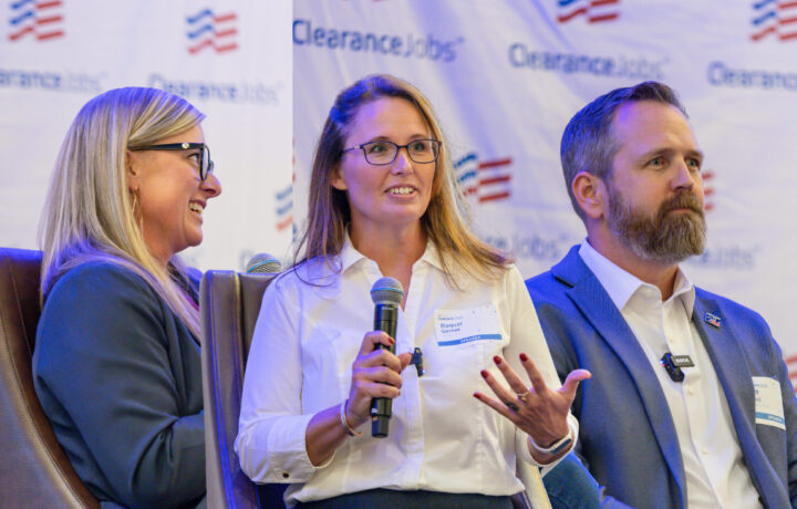 Three people are seated on a panel in front of a backdrop with the "ClearanceJobs" logo. The woman in the center, wearing glasses and a white shirt, is speaking into a microphone and gesturing with her hand. The woman on the left, also wearing glasses and a dark blazer, is smiling and looking toward the center speaker. The man on the right, with a beard and wearing a blue suit jacket, is looking forward attentively. All three have name tags and microphones clipped to their clothing.