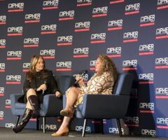 Two women are seated on stage in blue armchairs, engaged in a conversation. The woman on the left has long brown hair, wears a black outfit with knee-high shiny brown boots, and holds a microphone. The woman on the right has curly blonde hair, wears a patterned beige dress, and also holds a microphone. The backdrop behind them is dark blue with repeated logos and text reading "THE CIPHER BRIEF" and "Experts on National Security" in white and red.