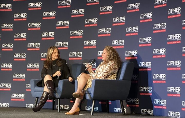 Two women are seated on stage in blue armchairs, engaged in a conversation. The woman on the left has long brown hair, wears a black outfit with knee-high shiny brown boots, and holds a microphone. The woman on the right has curly blonde hair, wears a patterned beige dress, and also holds a microphone. The backdrop behind them is dark blue with repeated logos and text reading "THE CIPHER BRIEF" and "Experts on National Security" in white and red.