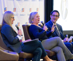 Three people are seated on stage in brown chairs, engaged in a panel discussion. The woman on the left has long blonde hair, wears glasses, a dark blazer, and holds a microphone and a notebook. The woman in the middle has short blonde hair, wears a dark long-sleeve top and gray pants, and is speaking into a microphone with a hand gesture. The man on the right wears glasses, a dark suit, white shirt, and tie, holding a microphone and some papers, smiling as he listens. The backdrop features a white banner with the "ClearanceJobs" logo.