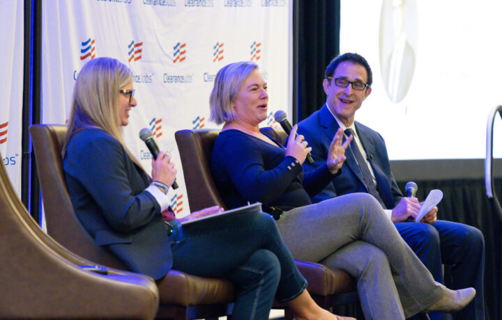 Three people are seated on stage in brown chairs, engaged in a panel discussion. The woman on the left has long blonde hair, wears glasses, a dark blazer, and holds a microphone and a notebook. The woman in the middle has short blonde hair, wears a dark long-sleeve top and gray pants, and is speaking into a microphone with a hand gesture. The man on the right wears glasses, a dark suit, white shirt, and tie, holding a microphone and some papers, smiling as he listens. The backdrop features a white banner with the "ClearanceJobs" logo.