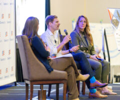 Three people are seated on stage in a panel discussion setting. The person in the middle, a man with short hair and a mustache, is speaking into a microphone and gesturing with his hand. He is wearing a white shirt and brown pants. To his right, a woman with long blonde hair, dressed in a dark blazer and pants, is holding a microphone and attentively listening. To his left, another woman with long brown hair, dressed in dark clothing, is also seated and facing the speaker. Behind them is a backdrop with the "ClearanceJobs" logo repeated, and to the right, a presentation board and a podium are visible.