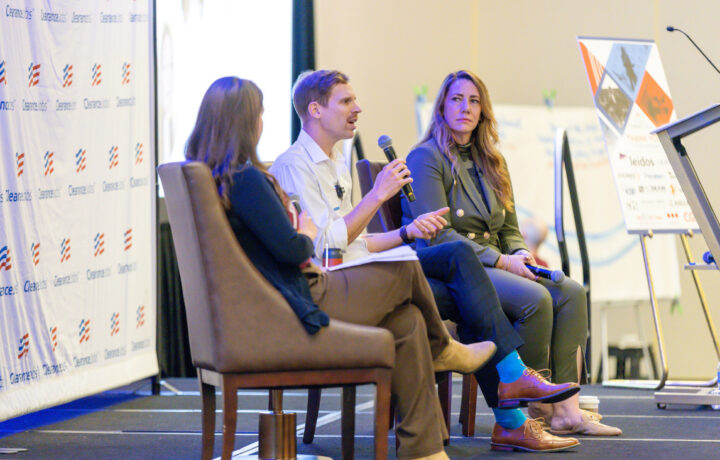 Three people are seated on stage in a panel discussion setting. The person in the middle, a man with short hair and a mustache, is speaking into a microphone and gesturing with his hand. He is wearing a white shirt and brown pants. To his right, a woman with long blonde hair, dressed in a dark blazer and pants, is holding a microphone and attentively listening. To his left, another woman with long brown hair, dressed in dark clothing, is also seated and facing the speaker. Behind them is a backdrop with the "ClearanceJobs" logo repeated, and to the right, a presentation board and a podium are visible.