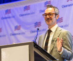A man with gray hair and a mustache, wearing brown glasses, a light gray suit, white shirt, and dark tie, is speaking at a podium with a microphone. The background features a banner with the "ClearanceJobs" logo and an American flag motif.