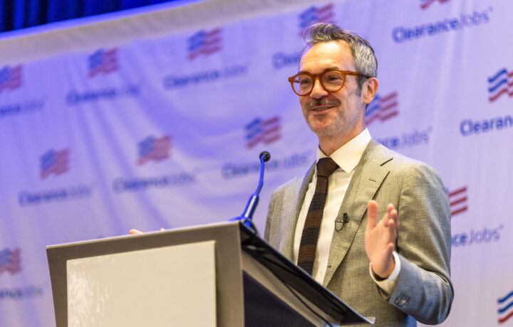 A man with gray hair and a mustache, wearing brown glasses, a light gray suit, white shirt, and dark tie, is speaking at a podium with a microphone. The background features a banner with the "ClearanceJobs" logo and an American flag motif.