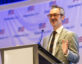 A man with gray hair and a mustache, wearing brown glasses, a light gray suit, white shirt, and dark tie, is speaking at a podium with a microphone. The background features a banner with the "ClearanceJobs" logo and an American flag motif.