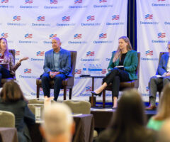 Four panelists sit on stage in front of a backdrop with repeated "ClearanceJobs" logos. The panelist on the far left, a woman with long hair wearing a leopard print jacket and black pants, is speaking into a microphone and gesturing with her hand. Next to her, a man in a blue blazer and light blue shirt sits with his hands clasped. The third panelist, a woman in a green blazer and black pants, holds a microphone and a notebook on her lap. The panelist on the far right, a man in a gray blazer and white shirt, looks down at a paper in his hands. In the foreground, the backs of audience members' heads are visible. A small table with water bottles is positioned between the second and third panelists.
