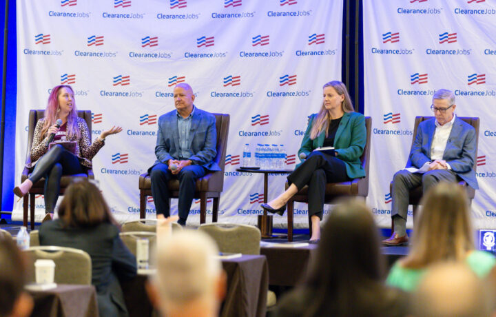 Four panelists sit on stage in front of a backdrop with repeated "ClearanceJobs" logos. The panelist on the far left, a woman with long hair wearing a leopard print jacket and black pants, is speaking into a microphone and gesturing with her hand. Next to her, a man in a blue blazer and light blue shirt sits with his hands clasped. The third panelist, a woman in a green blazer and black pants, holds a microphone and a notebook on her lap. The panelist on the far right, a man in a gray blazer and white shirt, looks down at a paper in his hands. In the foreground, the backs of audience members' heads are visible. A small table with water bottles is positioned between the second and third panelists.