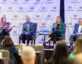 Four panelists sit on stage in front of a backdrop with repeated "ClearanceJobs" logos. The panelist on the far left, a woman with long hair wearing a leopard print jacket and black pants, is speaking into a microphone and gesturing with her hand. Next to her, a man in a blue blazer and light blue shirt sits with his hands clasped. The third panelist, a woman in a green blazer and black pants, holds a microphone and a notebook on her lap. The panelist on the far right, a man in a gray blazer and white shirt, looks down at a paper in his hands. In the foreground, the backs of audience members' heads are visible. A small table with water bottles is positioned between the second and third panelists.