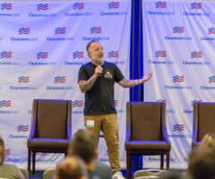A man with tattoos on both arms is standing on a stage, speaking into a microphone. He is wearing a black t-shirt with a "Mount Indie" logo and khaki pants. Behind him is a backdrop with repeated "ClearanceJobs" logos and American flag motifs. Four empty brown chairs are arranged on the stage, and an audience is visible in the foreground.