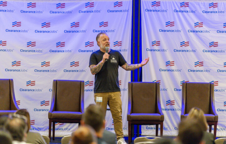 A man with tattoos on both arms is standing on a stage, speaking into a microphone. He is wearing a black t-shirt with a "Mount Indie" logo and khaki pants. Behind him is a backdrop with repeated "ClearanceJobs" logos and American flag motifs. Four empty brown chairs are arranged on the stage, and an audience is visible in the foreground.