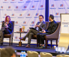 Three people are seated on a stage engaged in a panel discussion. The person on the left is a woman with long hair, holding a microphone and a tablet, wearing a dark blazer and sparkly pants. The two men on the right are seated next to each other; the man in the middle is speaking into a microphone, wearing glasses and a checked suit, while the man on the far right holds a microphone and notebook, dressed in a black suit with colorful socks. Behind them is a backdrop with the "ClearanceJobs" logo repeated, and to the right is a podium with a sign that reads "CONNECT 2025." A small table with awards and books is positioned between the woman and the man in the middle.