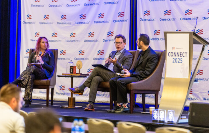 Three people are seated on a stage engaged in a panel discussion. The person on the left is a woman with long hair, holding a microphone and a tablet, wearing a dark blazer and sparkly pants. The two men on the right are seated next to each other; the man in the middle is speaking into a microphone, wearing glasses and a checked suit, while the man on the far right holds a microphone and notebook, dressed in a black suit with colorful socks. Behind them is a backdrop with the "ClearanceJobs" logo repeated, and to the right is a podium with a sign that reads "CONNECT 2025." A small table with awards and books is positioned between the woman and the man in the middle.