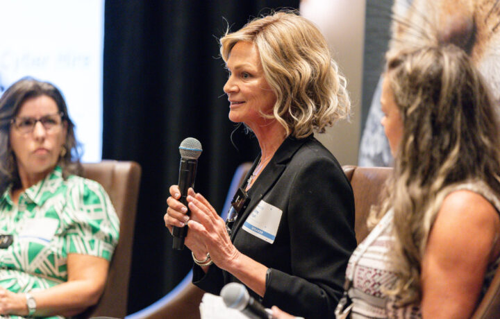 A woman with short blonde hair wearing a black blazer is speaking into a microphone during a panel discussion. She has a name tag on her blazer that reads "SPEAKER." Two other women are seated beside her, one in a green patterned shirt and glasses, and the other with long wavy hair wearing a sleeveless top. They are all seated in brown chairs.