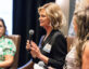 A woman with short blonde hair wearing a black blazer is speaking into a microphone during a panel discussion. She has a name tag on her blazer that reads "SPEAKER." Two other women are seated beside her, one in a green patterned shirt and glasses, and the other with long wavy hair wearing a sleeveless top. They are all seated in brown chairs.