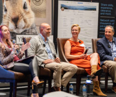 Four people are seated on stage chairs, engaged in a panel discussion. The woman on the left has long, wavy hair with purple highlights, wearing a leopard print jacket, black leggings, and brown high heels, holding a microphone and speaking. Next to her, a bald man in a light gray blazer, blue shirt, and beige pants is smiling, holding a microphone. The woman beside him wears an orange sleeveless dress and tan boots, smiling while holding a microphone. The man on the far right is dressed in a navy blazer, striped shirt, and beige pants, holding a microphone and smiling. Behind them are large posters, one featuring a close-up of a chipmunk and another with text about sharing skills and networking. Bottles of water are on the floor near their feet.