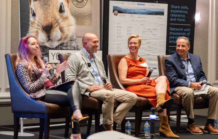 Four people are seated on stage chairs, engaged in a panel discussion. The woman on the left has long, wavy hair with purple highlights, wearing a leopard print jacket, black leggings, and brown high heels, holding a microphone and speaking. Next to her, a bald man in a light gray blazer, blue shirt, and beige pants is smiling, holding a microphone. The woman beside him wears an orange sleeveless dress and tan boots, smiling while holding a microphone. The man on the far right is dressed in a navy blazer, striped shirt, and beige pants, holding a microphone and smiling. Behind them are large posters, one featuring a close-up of a chipmunk and another with text about sharing skills and networking. Bottles of water are on the floor near their feet.