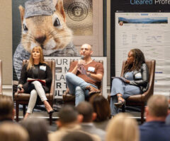 Three panelists are seated on stage in brown leather chairs, engaged in a discussion. The panelist in the center, a bald man wearing glasses, a brown shirt, and light blue jeans, is speaking into a microphone. To his left, a woman with long blonde hair, dressed in a black top and white pants, holds a microphone and a notebook. To his right, a woman with long braided hair, wearing a black leather jacket and light blue pants, holds papers. Behind them is a large banner featuring a close-up image of a squirrel in military uniform and text promoting professional networking and clearance. The audience is visible in the foreground, facing the panelists.