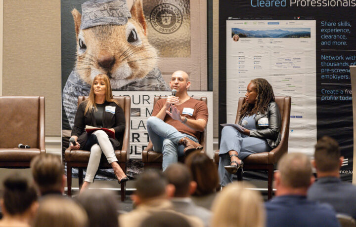 Three panelists are seated on stage in brown leather chairs, engaged in a discussion. The panelist in the center, a bald man wearing glasses, a brown shirt, and light blue jeans, is speaking into a microphone. To his left, a woman with long blonde hair, dressed in a black top and white pants, holds a microphone and a notebook. To his right, a woman with long braided hair, wearing a black leather jacket and light blue pants, holds papers. Behind them is a large banner featuring a close-up image of a squirrel in military uniform and text promoting professional networking and clearance. The audience is visible in the foreground, facing the panelists.