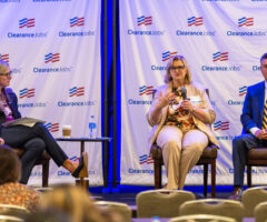 Three panelists sit on stage in front of a backdrop with the ClearanceJobs logo. The woman on the left wears glasses, a black blazer, and black pants, holding a microphone and a folder. The woman in the center wears glasses, a beige suit, and a floral blouse, speaking into a microphone. The man on the right wears a dark suit with a white shirt and a yellow-striped tie, holding a microphone. A small table with a water bottle and a coffee cup is between the left and center panelists. Audience members are partially visible in the foreground.