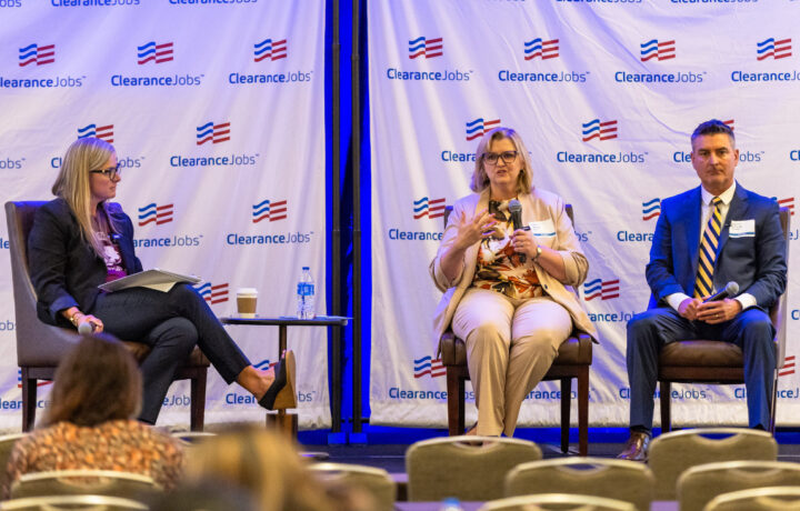 Three panelists sit on stage in front of a backdrop with the ClearanceJobs logo. The woman on the left wears glasses, a black blazer, and black pants, holding a microphone and a folder. The woman in the center wears glasses, a beige suit, and a floral blouse, speaking into a microphone. The man on the right wears a dark suit with a white shirt and a yellow-striped tie, holding a microphone. A small table with a water bottle and a coffee cup is between the left and center panelists. Audience members are partially visible in the foreground.