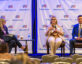 Three panelists sit on stage in front of a backdrop with the ClearanceJobs logo. The woman on the left wears glasses, a black blazer, and black pants, holding a microphone and a folder. The woman in the center wears glasses, a beige suit, and a floral blouse, speaking into a microphone. The man on the right wears a dark suit with a white shirt and a yellow-striped tie, holding a microphone. A small table with a water bottle and a coffee cup is between the left and center panelists. Audience members are partially visible in the foreground.