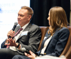 A man in a gray suit and pink tie is speaking into a microphone while seated. He has a name tag labeled "SPEAKER." Next to him, a woman with long brown hair, wearing a navy blazer and white top, is holding a microphone and looking at him. They are seated on brown chairs with a blurred presentation screen in the background.