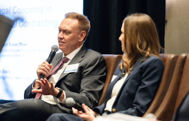 A man in a gray suit and pink tie is speaking into a microphone while seated. He has a name tag labeled "SPEAKER." Next to him, a woman with long brown hair, wearing a navy blazer and white top, is holding a microphone and looking at him. They are seated on brown chairs with a blurred presentation screen in the background.
