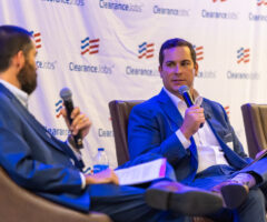 Two men in suits are seated on stage chairs, each holding a microphone and papers, engaged in a discussion. The backdrop features a repeated logo with red and blue stripes and the text "ClearanceJobs." One man has a beard and is facing the other, who is clean-shaven and looking attentively at his counterpart.