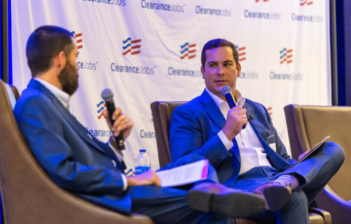 Two men in suits are seated on stage chairs, each holding a microphone and papers, engaged in a discussion. The backdrop features a repeated logo with red and blue stripes and the text "ClearanceJobs." One man has a beard and is facing the other, who is clean-shaven and looking attentively at his counterpart.