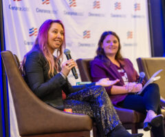 Two women are seated on brown chairs on a stage, engaged in a discussion. The woman on the left has long pink hair, wears a black blazer and sparkly black pants, and is holding a microphone while speaking. The woman on the right has long brown hair, wears a purple blazer and black pants, and holds a microphone and some papers. The background features a white banner with the "ClearanceJobs" logo repeated across it.
