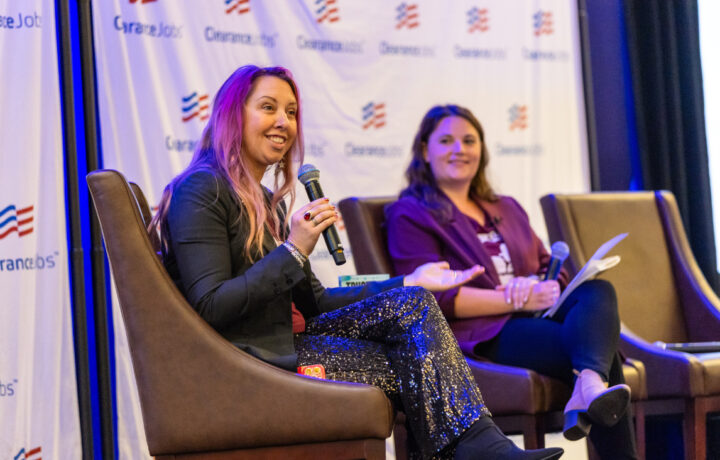 Two women are seated on brown chairs on a stage, engaged in a discussion. The woman on the left has long pink hair, wears a black blazer and sparkly black pants, and is holding a microphone while speaking. The woman on the right has long brown hair, wears a purple blazer and black pants, and holds a microphone and some papers. The background features a white banner with the "ClearanceJobs" logo repeated across it.