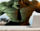 A muscular green Hulk character wearing black glasses and a black necktie, sitting behind a desk with a laptop in a blurred office background.