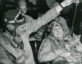 Two men in vintage pilot gear, including leather helmets and goggles, are interacting with a cat. One man is holding the cat while the other dangles a string above it, engaging the cat's attention. Both men appear to be smiling and seated in a room with other people in the background.