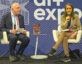 A speaker participates in a discussion at the AI+ Expo while seated on stage beside an interviewer. A copy of Project Maven: A Marine Colonel, His Team, and the Dawn of AI Warfare by Katrina Manson is displayed on a small table between them. The event backdrop features AI+ Expo branding, highlighting a conversation about artificial intelligence and national security.